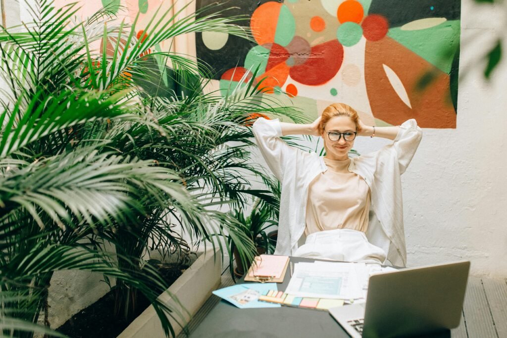 Uma mulher sorridente segura documentos diante de um mural colorido e moderno, com plantas ao redor, transmitindo leveza, organização e um ambiente profissional descontraído.